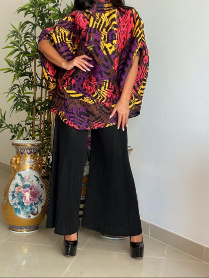 Woman wearing a colorful patterned top and black pants standing indoors with a plant and vase in the background.
